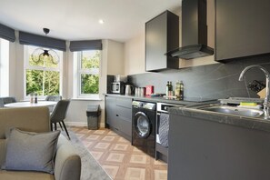 A compact kitchen with a built-in washing machine, a dining area with a table and chairs, and large windows providing natural light. - The Olive Tree | Superior 2-Bed Apartment (Kingston upon Hull)