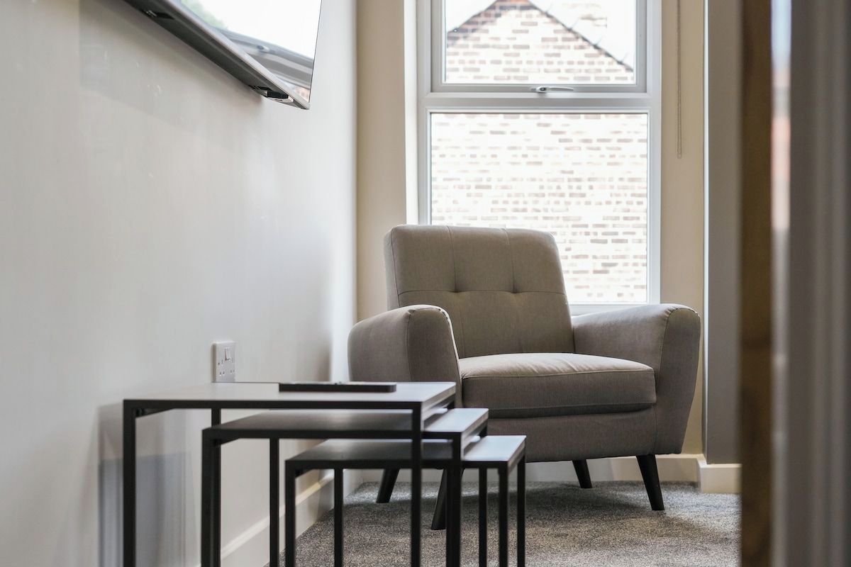 A corner room with a window, a grey armchair, and a small table.