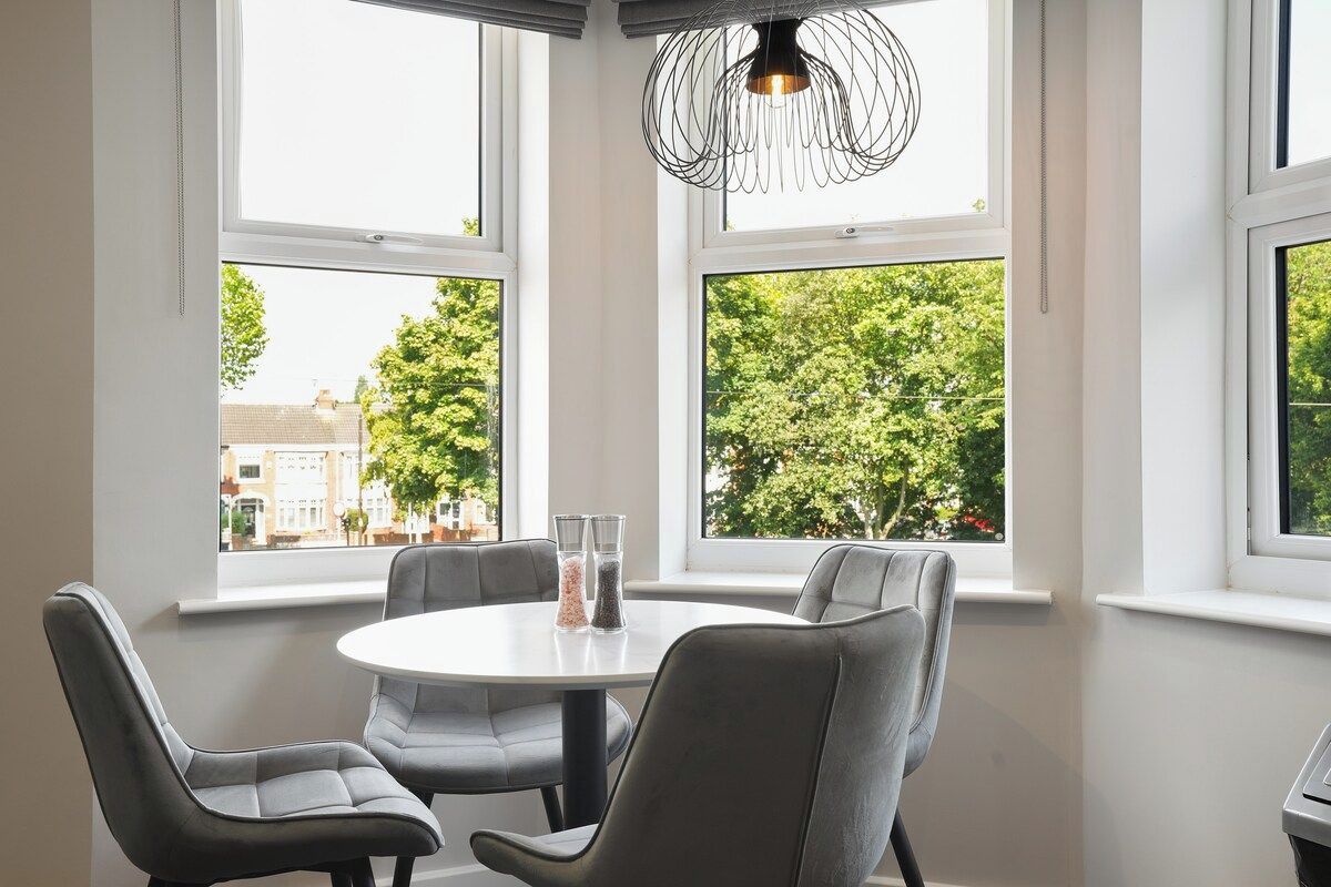 A dining area with a round table and four chairs, large windows with a view of trees and buildings, and a unique pendant light fixture.