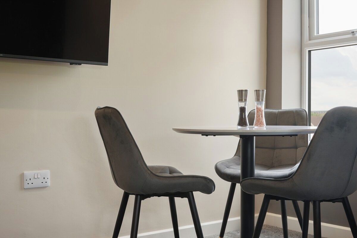 A room with a flat-screen TV mounted on the wall, a round table with two chairs, and a window providing a view.