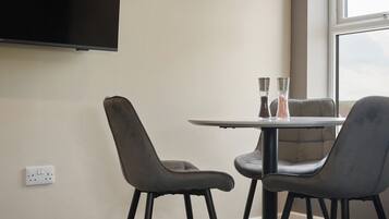 A room with a flat-screen TV mounted on the wall, a round table with two chairs, and a window providing a view.