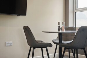 A room with a flat-screen TV mounted on the wall, a round table with two chairs, and a window providing a view. - The Olive Tree | Stylish 2-Bed Loft (Kingston upon Hull)