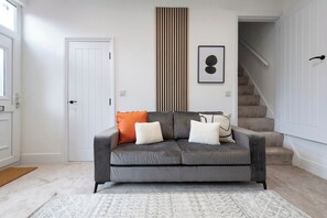 Ein neutral eingerichtetes Wohnzimmer mit grauem Sofa, einem gemusterten Teppich, einer vertikalen Holzlamelle und einem gerahmten Bild an der Wand.