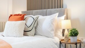Una cama bien tendida con varios almohadones, una lámpara de noche y una planta pequeña sobre una mesita.