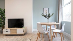 Una sala de estar moderna con un televisor de pantalla plana, un aparador de madera, una mesa de comedor redonda con sillas blancas, una planta en maceta y un cuadro en la pared.
