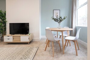Ein modernes Wohnzimmer mit einem Flachbildfernseher, einer Holzkommode, einem runden Esstisch mit weißen Stühlen, einem Topfpflanzen und einem gerahmten Bild an der Wand.