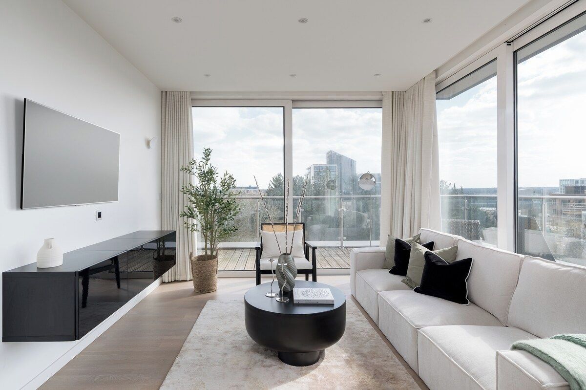 A modern living room with a flat-screen TV, a black coffee table, a white sofa, and a large window offering a city view.