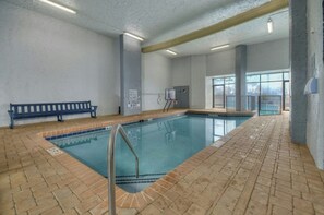 An indoor swimming pool with a bench, safety signs, and a view of the city through large windows. - Bluewater Resort 920-Oceanfront I/O Pool-Hot tub (Myrtle Beach)