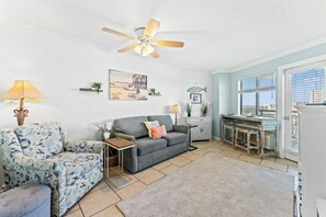 A living room with a grey sofa, a patterned armchair, a ceiling fan, and a view of the city through a window. - Bluewater Resort 920-Oceanfront I/O Pool-Hot tub (Myrtle Beach)