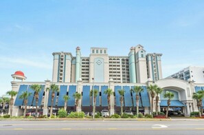 A multi-story hotel with a central clock tower, palm trees, and a parking lot. - Compass Cove Mariner 1020- 3BR Great Amenities (Myrtle Beach)