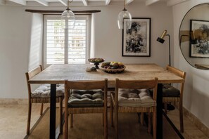A dining area with a wooden table, six chairs with cushions, a fruit bowl, and two pendant lights. - Rose Cottage (Helston)