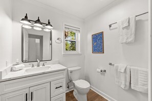 Ein Badezimmer mit weißem Waschtisch, Toilette, einem Fenster mit Blick auf Grünflächen und einem gerahmten Kunstwerk an der Wand.