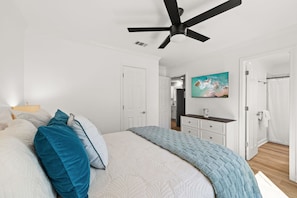 Quarto com cama, ventilador de teto, um quadro e banheiro visível através de uma porta aberta.