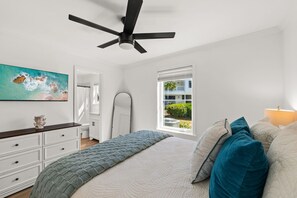 Ein Schlafzimmer mit einem Bett, einem Deckenventilator, einem Fenster mit Blick und einem Schminktisch. An der Wand hängt ein Gemälde.