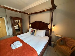 Quarto de hotel com uma cama grande, cabeceira de madeira, espelho, cadeira de vime e uma pequena mesa de cabeceira.