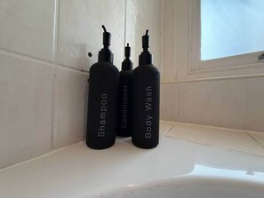 Drei schwarze Flaschen mit den Aufschriften 'Shampoo', 'Spülung' und 'Duschgel' auf einer weißen Waschtischplatte.