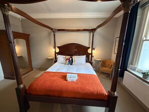 Ein Himmelbett mit weißer Bettdecke und orangefarbener Tagesdecke, begleitet von zwei Wandlampen und einem Spiegel.