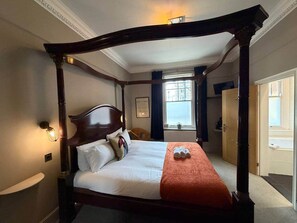 Uma cama de dossel com roupa de cama branca e um cobertor laranja, uma mesa de cabeceira com abajur e uma janela com cortinas.