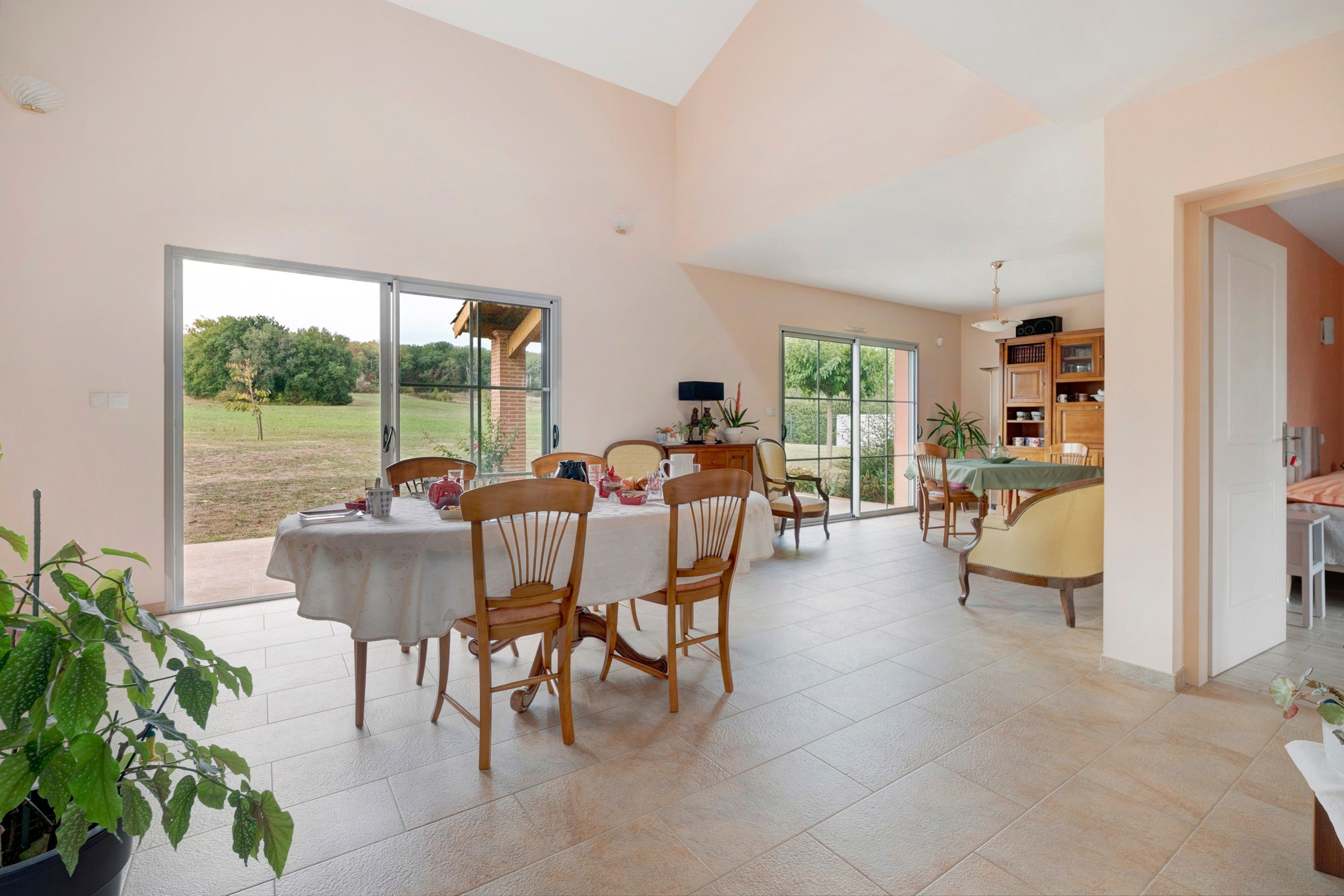 Un vaste espace de salle à manger avec une grande table et des chaises, un coin cuisine doté de placards et d’une table, et une vue sur un jardin grâce à de grandes portes-fenêtres coulissantes.