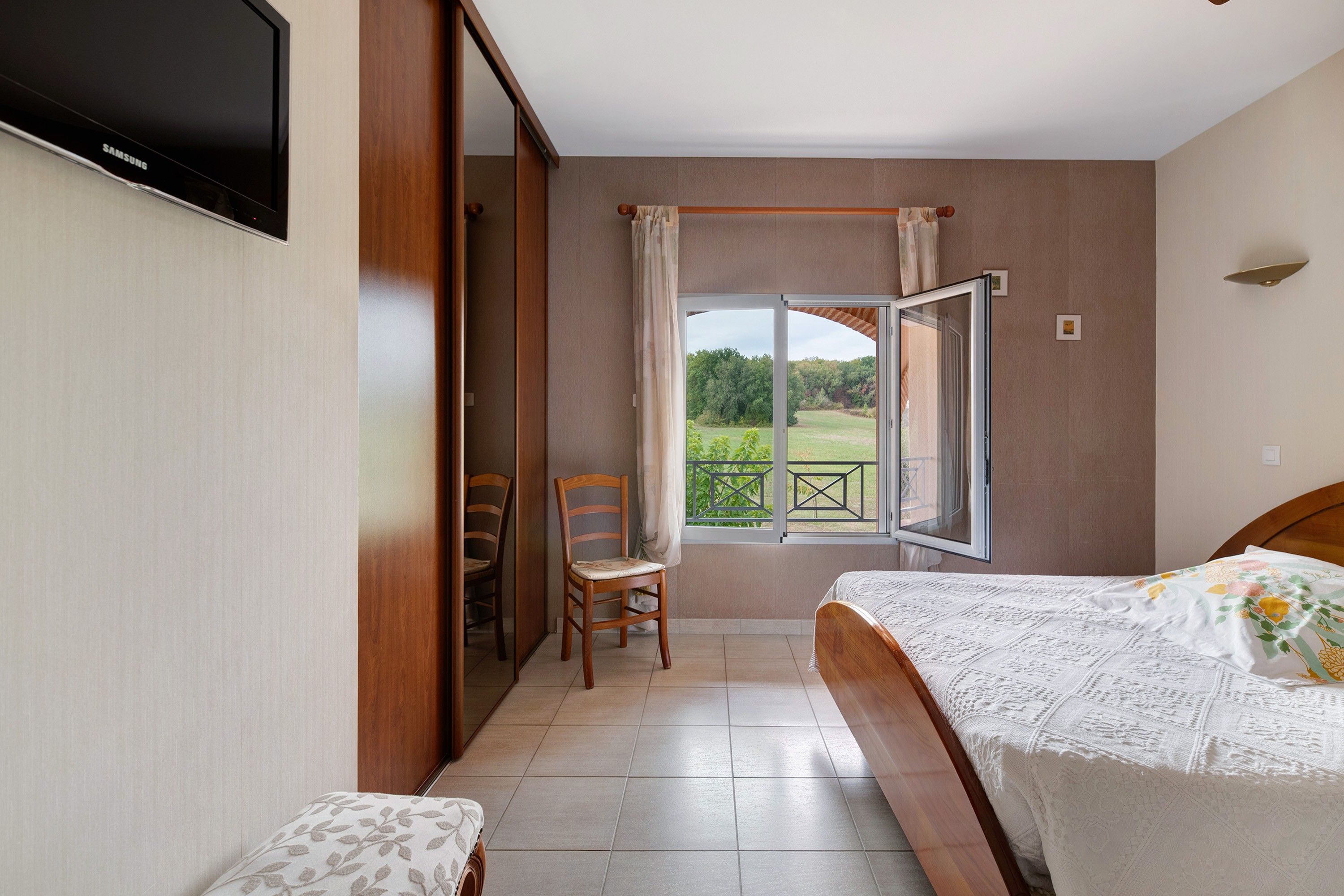 A bedroom with a bed, a chair, a TV, and a window with a view of greenery.