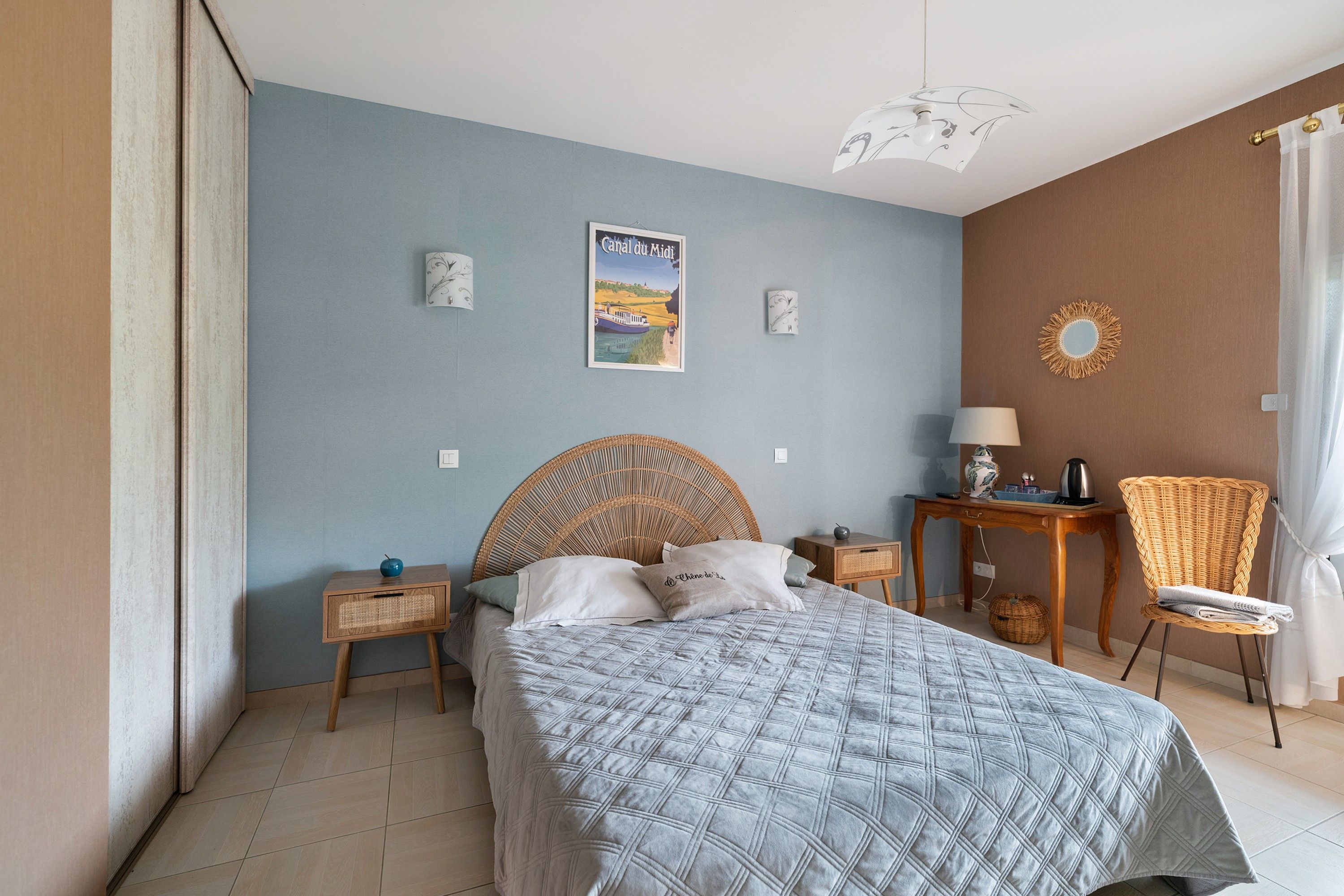 Een slaapkamer met een bed, nachtkastje, stoel, tafel en een muur met een afbeelding.
