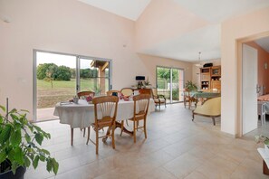 A spacious dining area with a large table and chairs, a kitchen area with cabinets and a dining table, and a view of a garden through large sliding glass doors.