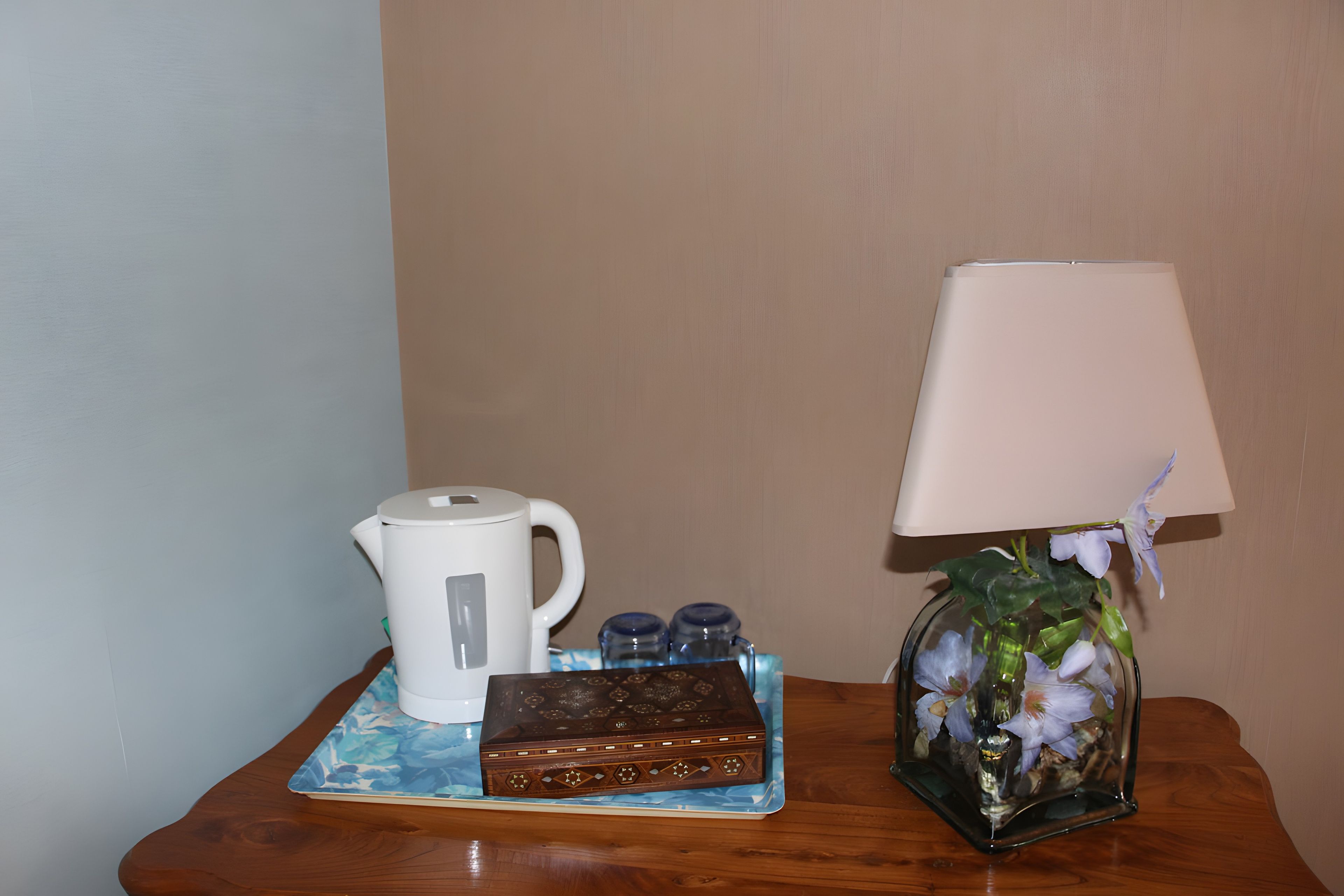 Une table de chevet avec une bouilloire blanche, une boîte décorative, un vase en verre avec des fleurs et une lampe.