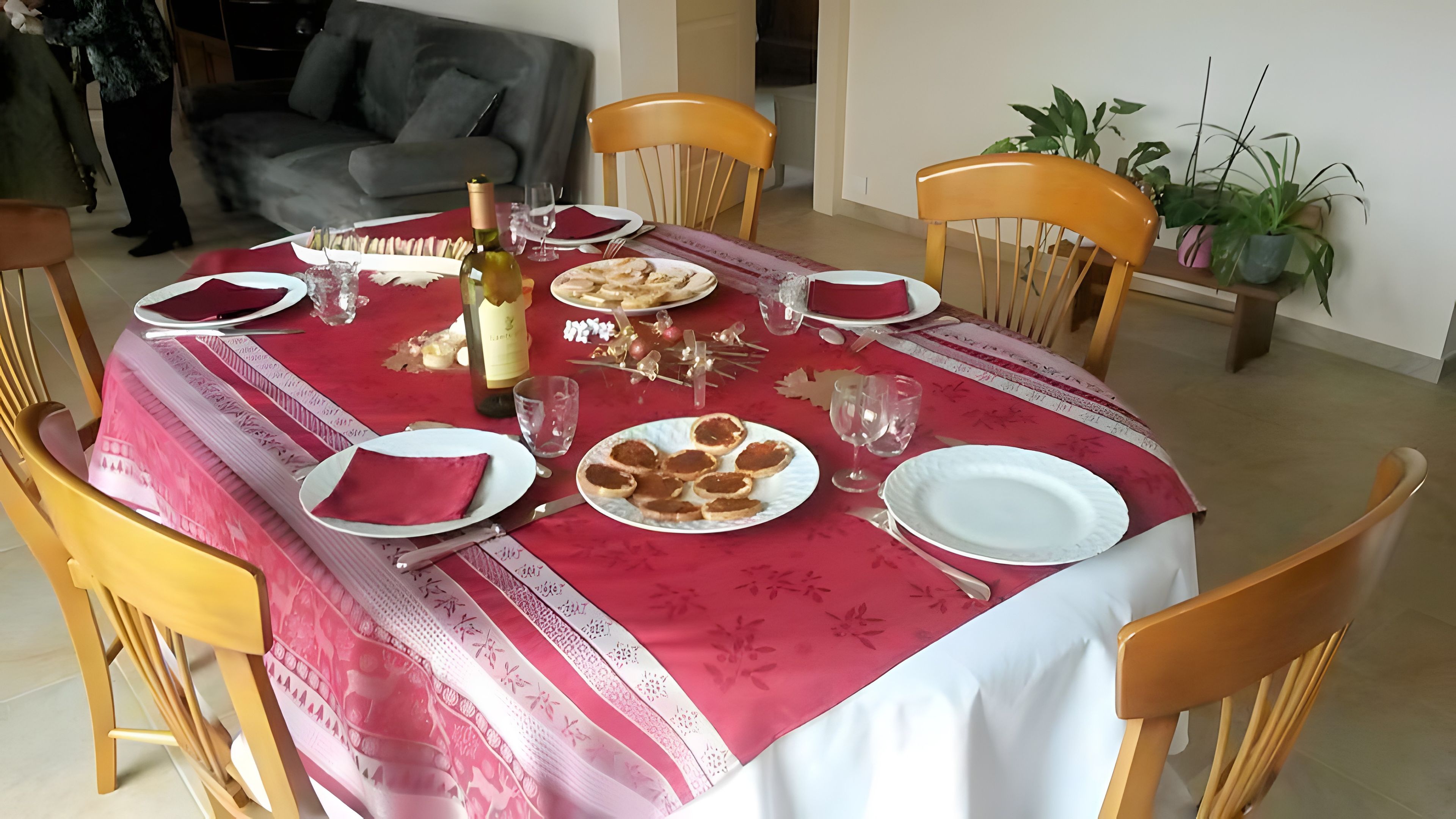 Ein Esstisch für vier Personen, gedeckt mit einer roten Tischdecke, Tellern, Gläsern und einer Weinflasche.