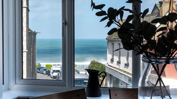 Ein Zimmer mit Meerblick durch ein großes Fenster, eine Küstpflanze und eine schwarze Karaffe auf dem Fensterbrett.