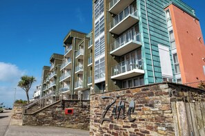 A multi-story residential building with balconies, a stone wall with a metal sign, and a clear blue sky. - Luxury Fistral Beach Apartment Newquay, Cornwall Parking (Cornwall)