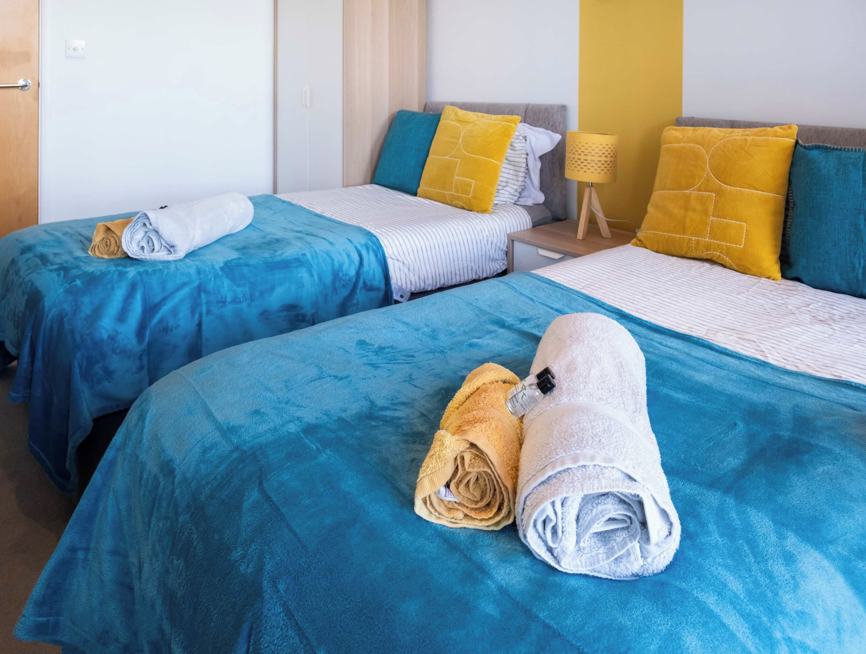 A hotel room with two beds, each with a blue bedspread and a rolled towel. There is a bedside table with a lamp and a small table lamp.