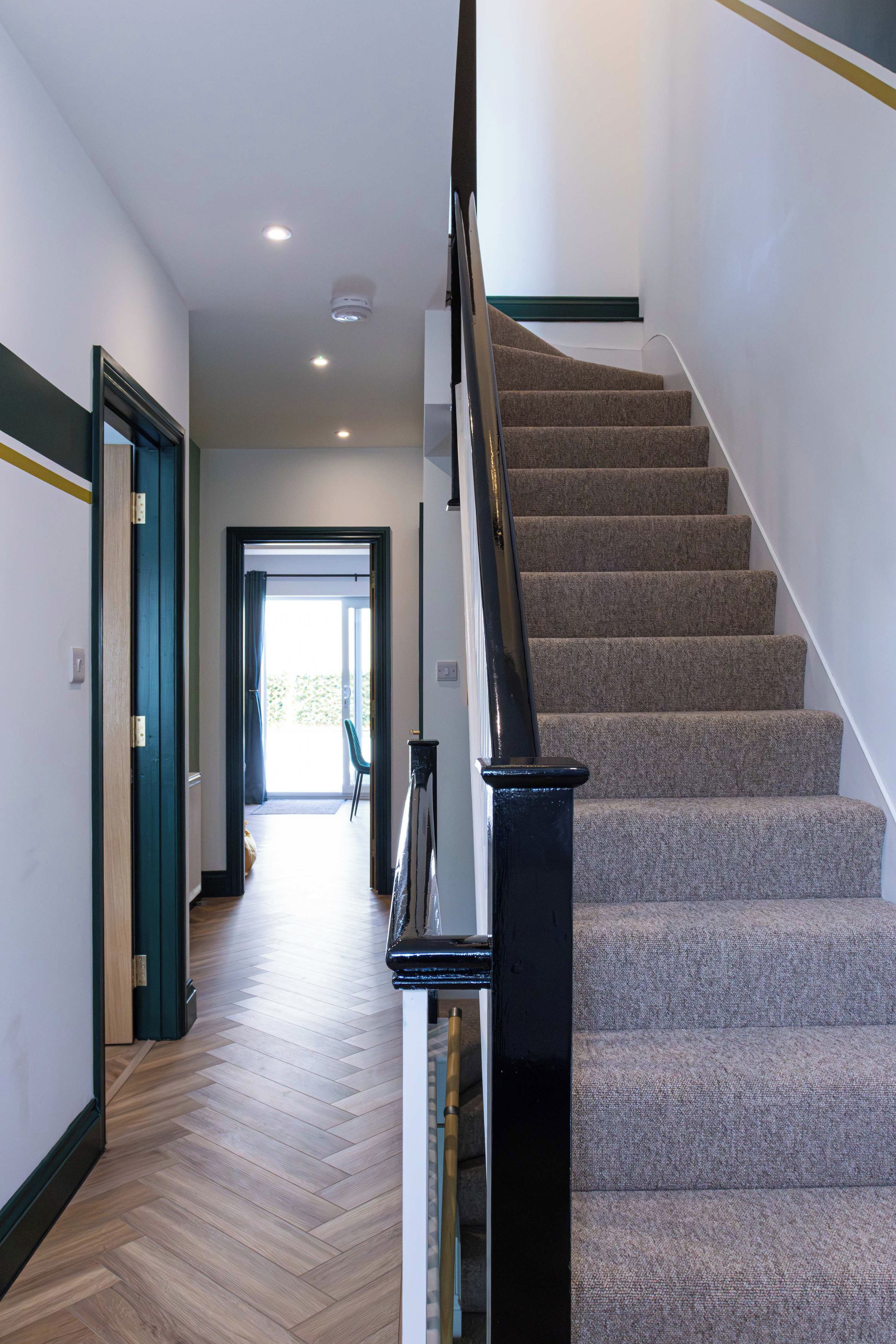 A modern interior with a carpeted staircase, wooden flooring, and a view into a bright room.