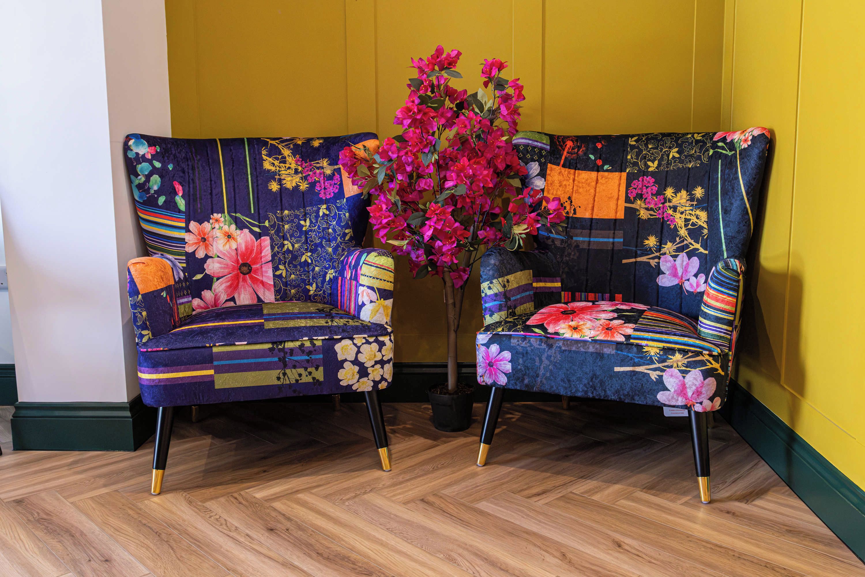 Two upholstered chairs with floral upholstery and black metal legs, positioned in front of a yellow wall and a potted plant with pink flowers.