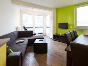 A modern living room with a sofa, coffee table, and a dining area with chairs. There is a television on the wall and a window providing natural light. - Holiday Apartment With 2 Bedrooms (Norderney)