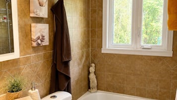 Une salle de bain équipée d’une baignoire, de toilettes, d’un lavabo et d’une fenêtre donnant sur un espace verdoyant.