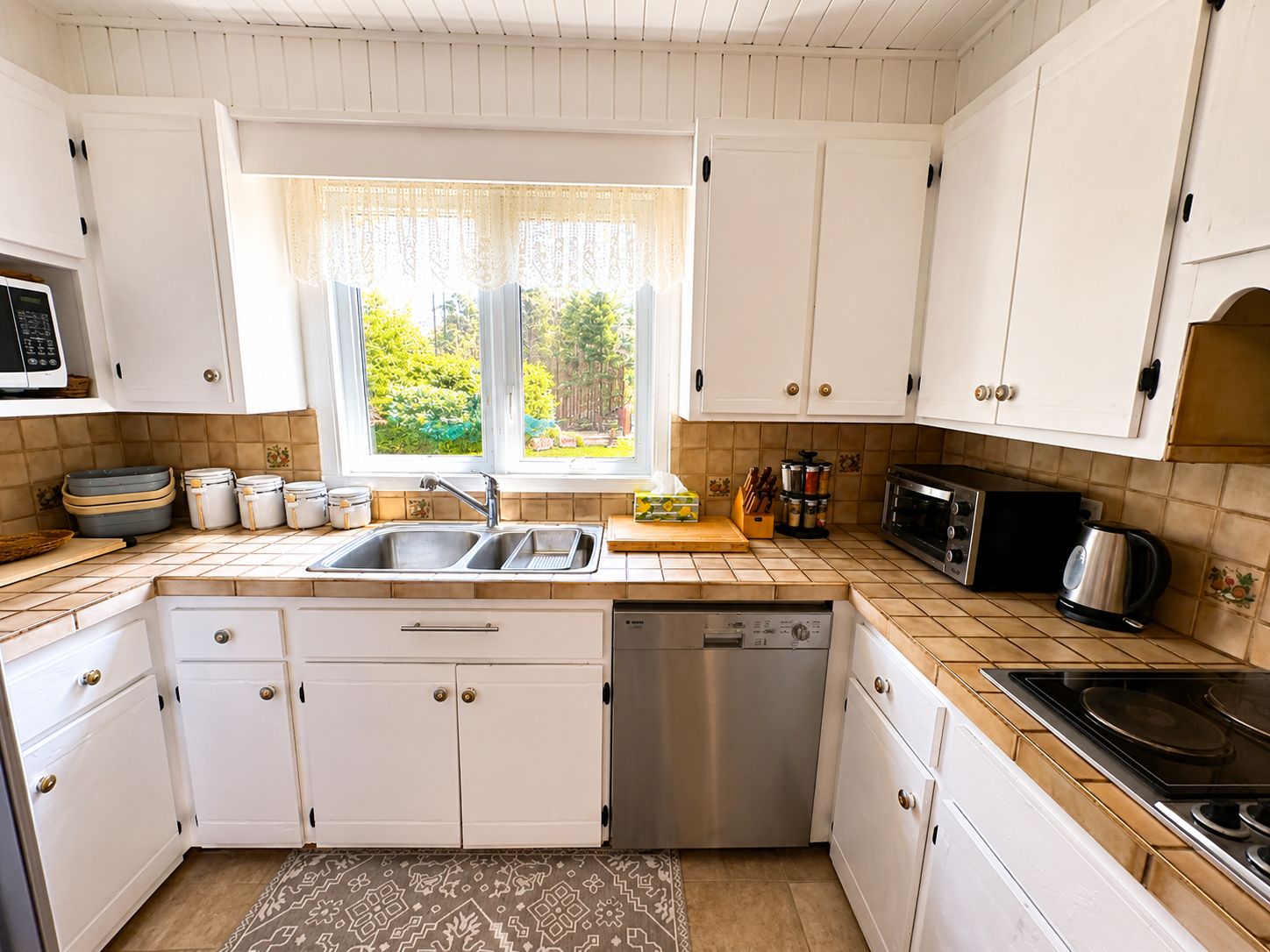 Une cuisine avec des meubles blancs, un évier, un lave-vaisselle, une cuisinière et une fenêtre donnant sur un paysage verdoyant.