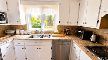 Une cuisine avec des meubles blancs, un évier, un lave-vaisselle, une cuisinière et une fenêtre donnant sur un paysage verdoyant.
