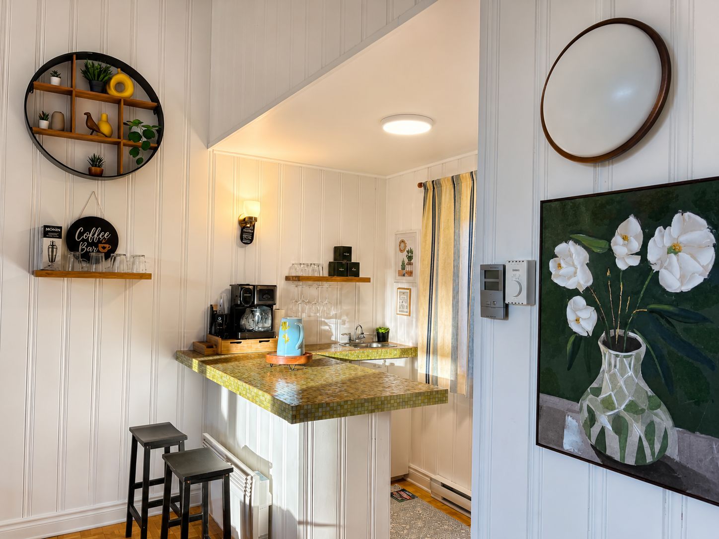 A kitchen with a coffee bar, a round shelf with plants, a painting of flowers, and a tiled backsplash.