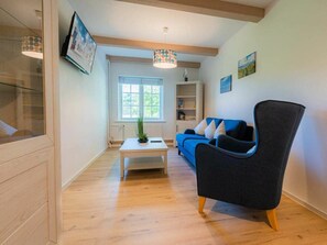 Ein modernes Wohnzimmer mit Holzboden, einem blauen Sofa, einem dunklen Sessel, einem weißen Couchtisch und einem Fenster mit Blick auf Grünflächen.