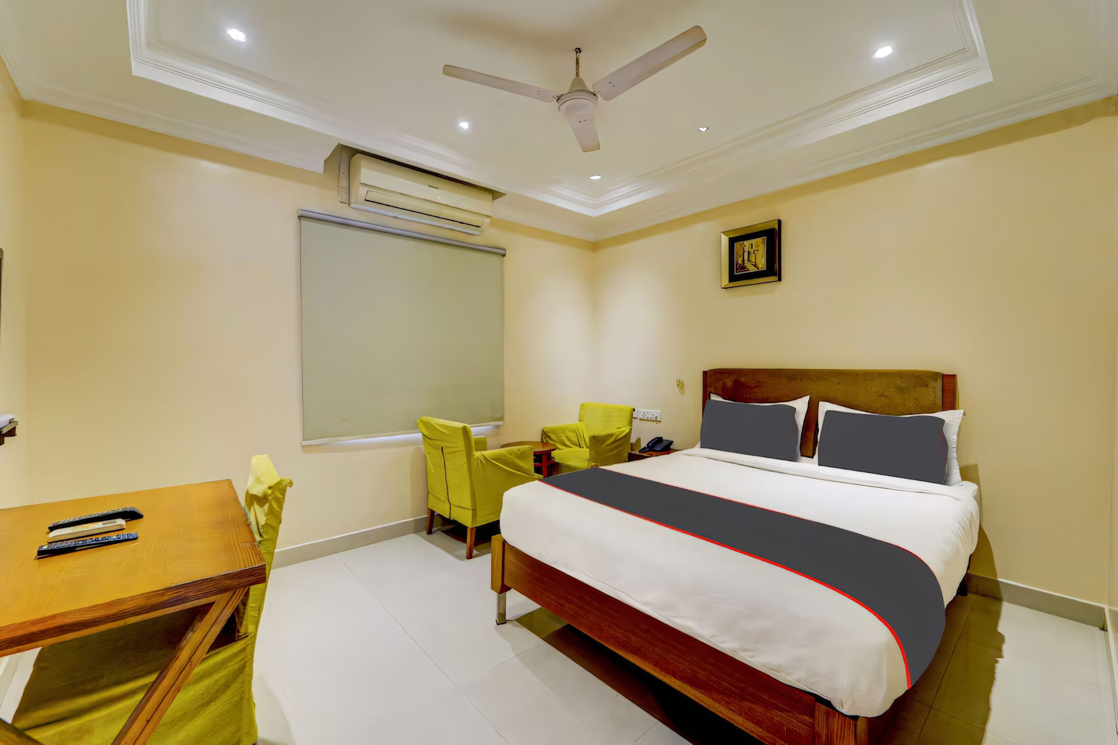 Habitación de hotel con cama, escritorio, silla, ventilador de techo, equipo de aire acondicionado y un cuadro colgado en la pared.