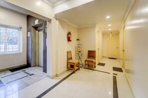 A hotel hallway with a fire extinguisher, two wooden chairs, a water dispenser, and a door at the end.