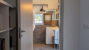 Un pasillo estrecho que conduce a una cocina con ventana, un radiador y un taburete pequeño.