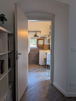 Een smalle gang die uitkomt in een keuken met een raam, een radiator en een klein krukje.