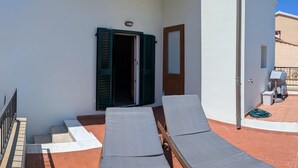 Un balcón con dos sillones de descanso, una puerta con contraventanas verdes y una mesita.