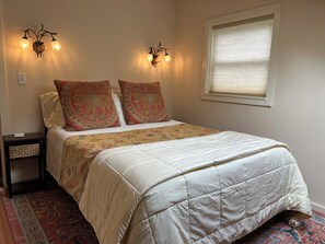 Un dormitorio con una cama, dos luces fijadas a la pared, un ventanal con persianas y una mesita de noche con lámpara.