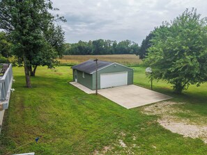 A green metal shed with a white garage door, a concrete driveway, and a grassy yard with trees.