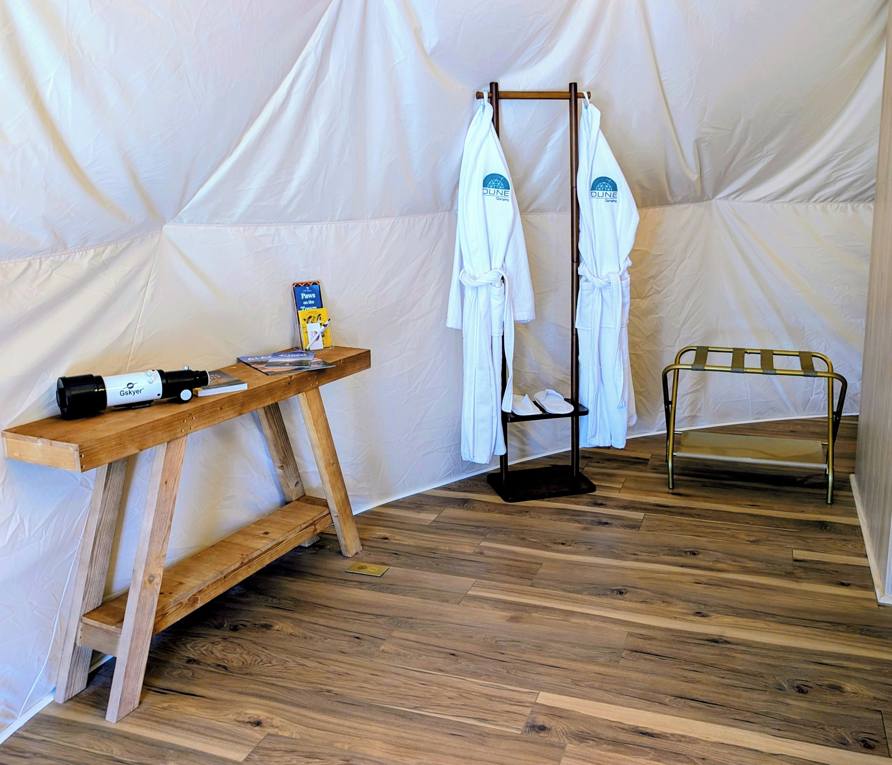 Interior de uma tenda com uma mesa de madeira, um porta-toalhas e uma cama portátil.