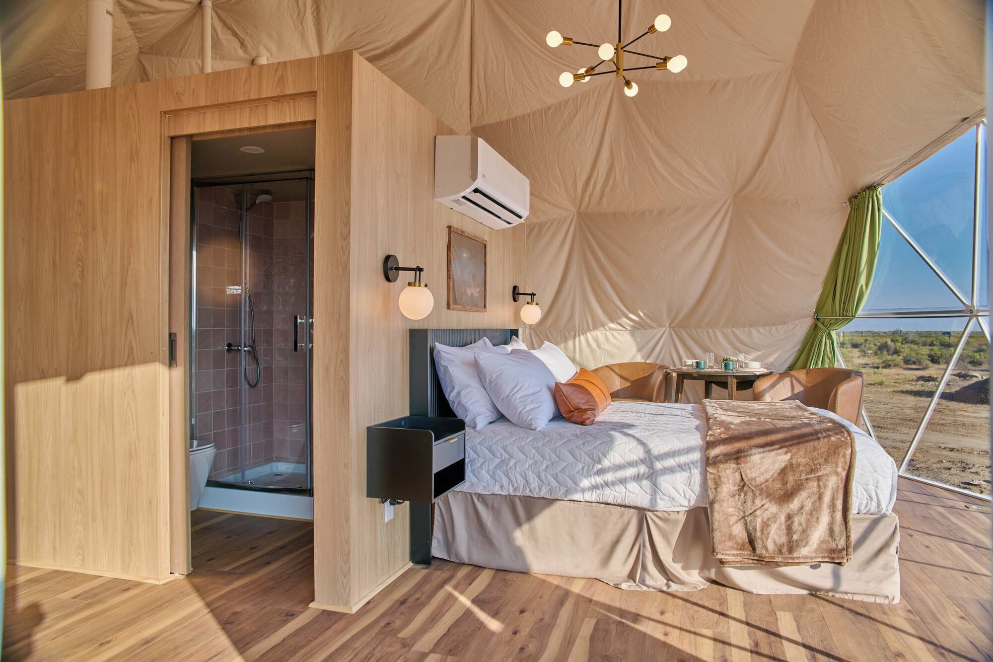 A tented accommodation with a bed, bedside table, lamp, and a view of the desert.