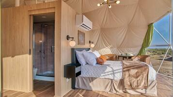 A tented accommodation with a bed, bedside table, lamp, and a view of the desert.