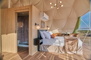 A tented accommodation with a bed, bedside table, lamp, and a view of the desert.